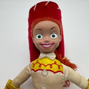 May include: A Jessie doll from the Toy Story franchise. The doll has a red hat, orange hair, and a yellow and cream-colored shirt with a red trim. The doll's blue jeans and yellow wrist cuffs are also visible.