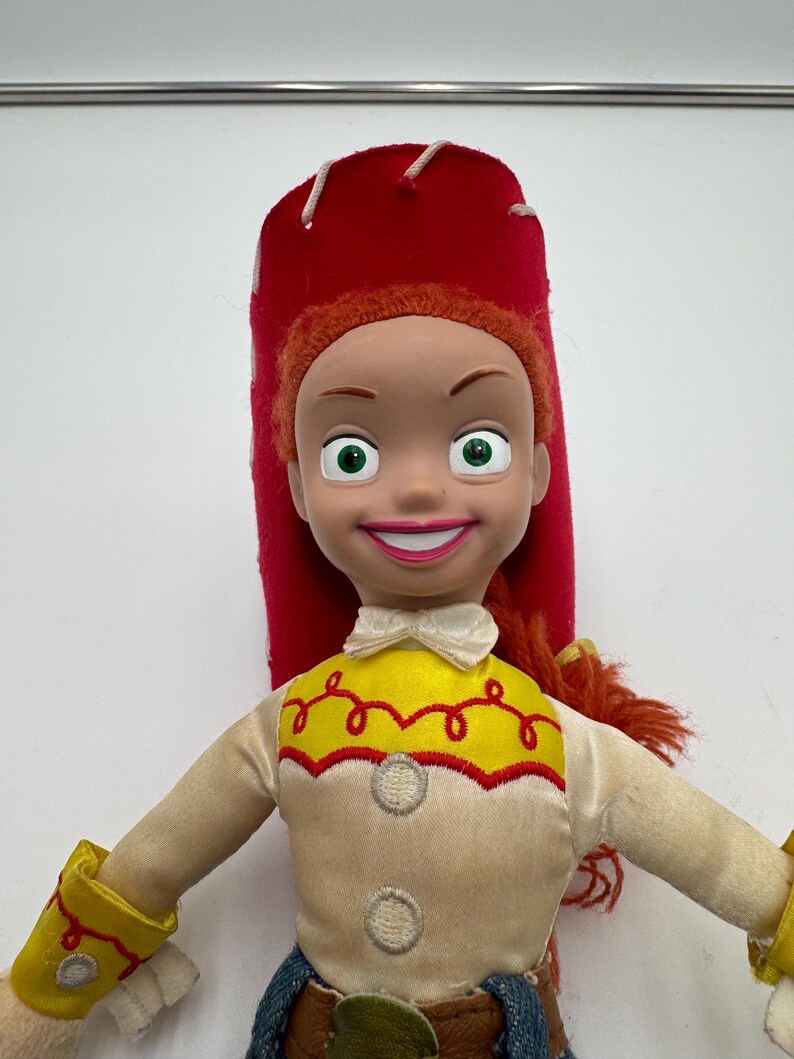 May include: A Jessie doll from the Toy Story franchise. The doll has a red hat, orange hair, and a yellow and cream-colored shirt with a red trim. The doll's blue jeans and yellow wrist cuffs are also visible.