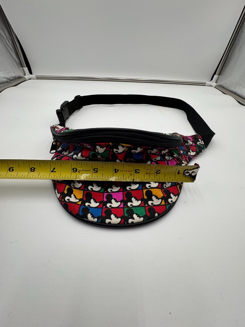 May include: A colorful fanny pack featuring a repeating grid pattern of Mickey Mouse heads in red, blue, yellow, and green squares. A yellow measuring tape is placed across the front, showing a width of approximately 8 inches for the bag.