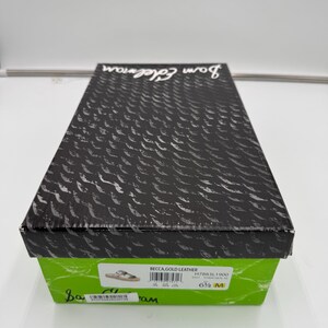 May include: A rectangular shoe box with a black lid featuring a textured pattern and the brand name "Sam Edelman." The box has a green base with product information, including "BECCA, GOLD LEATHER" and size 6 1/2 M.
