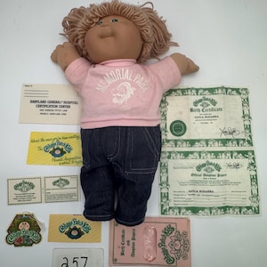Puede incluir: Una muñeca Cabbage Patch Kids vintage con una camiseta rosa con el texto "Memorial Park" y pantalones vaqueros azul oscuro. Incluye certificados de nacimiento, papeles de adopción y pegatinas de Cabbage Patch Kids. Cabello castaño claro.