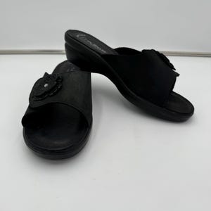 May include: Two black slide sandals with a small wedge heel. Each sandal features a decorative floral accent on the strap. The sandals are made of a matte material and are set against a white background.
