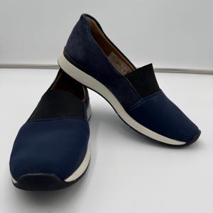 以下が含まれることがあります： ネイビーブルーのスリッポンシューズ。上部に黒いゴムバンドが付いています。靴は白いソールと、ダークブルーのスエードのような素材のかかとが特徴です。快適さと履きやすさを追求したデザインです。
