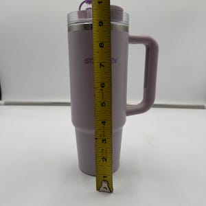 Op de afbeelding: Een lavendelkleurige Stanley reismok met een handvat en een doorzichtig deksel. De mok is ongeveer 25 cm hoog. Op de zijkant van de mok staat het woord "Stanley". Het deksel heeft een kleine paarse dop.