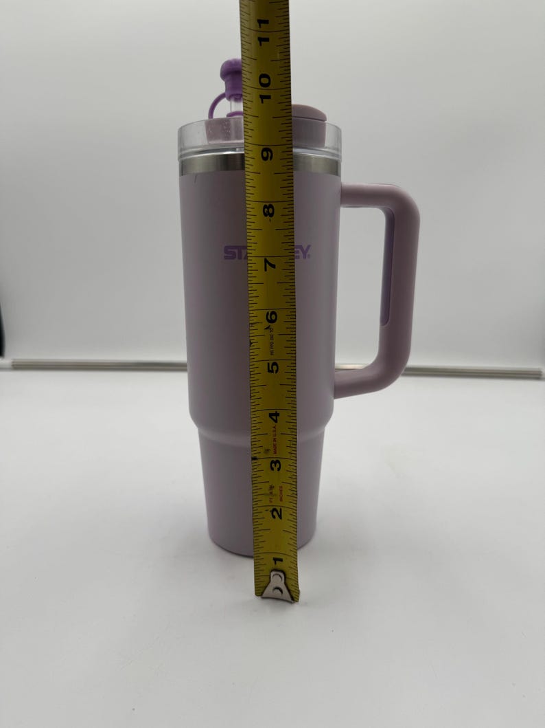 Op de afbeelding: Een lavendelkleurige Stanley reismok met een handvat en een doorzichtig deksel. De mok is ongeveer 25 cm hoog. Op de zijkant van de mok staat het woord "Stanley". Het deksel heeft een kleine paarse dop.