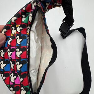 May include: A fanny pack featuring a repeating Mickey Mouse pattern set against a grid of vibrant red, blue, green, and pink squares. The bag has a black zipper closure, a white interior lining, and an adjustable black nylon strap with a plastic buckle.