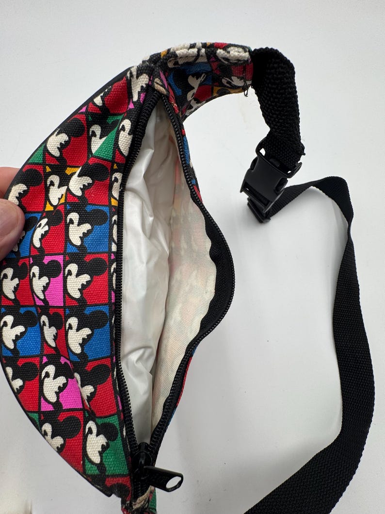 May include: A fanny pack featuring a repeating Mickey Mouse pattern set against a grid of vibrant red, blue, green, and pink squares. The bag has a black zipper closure, a white interior lining, and an adjustable black nylon strap with a plastic buckle.