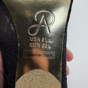 K&ouml;nnte beinhalten: Nahaufnahme der Sohle eines schwarzen Spitzen-High-Heel-Schuhs. Die goldene Sohle zeigt das Markenlogo, Gr&ouml;&szlig;enangaben (USA 8 1/2 M, EUR 39 1/2) und den Text "Leather Sock".