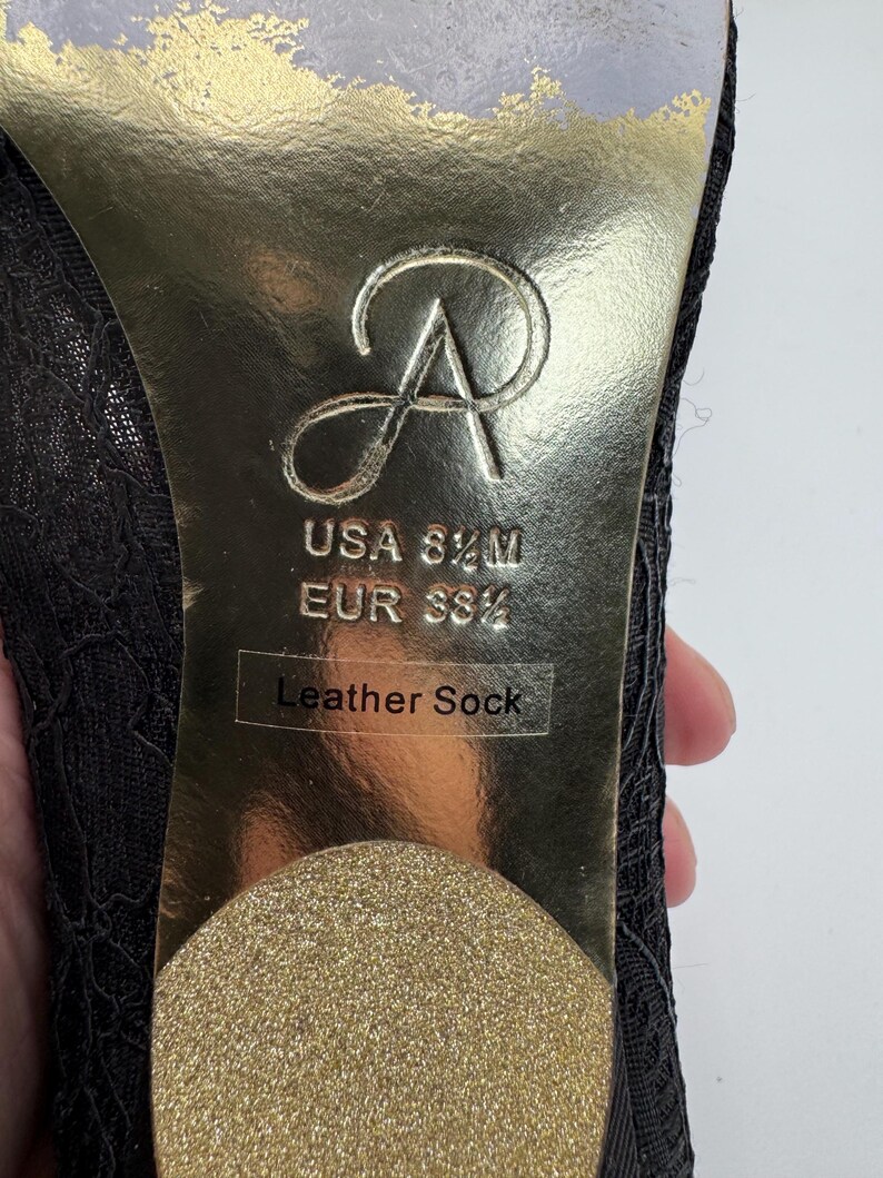 K&ouml;nnte beinhalten: Nahaufnahme der Sohle eines schwarzen Spitzen-High-Heel-Schuhs. Die goldene Sohle zeigt das Markenlogo, Gr&ouml;&szlig;enangaben (USA 8 1/2 M, EUR 39 1/2) und den Text "Leather Sock".
