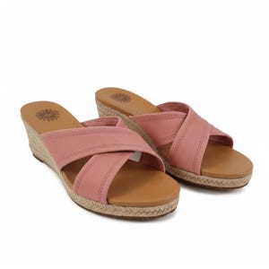 Puede incluir: Un par de sandalias de cuña rosas con correas cruzadas. Las sandalias tienen una plantilla marrón y una suela de yute tejida. El logotipo de UGG es visible en la plantilla. Sobre un fondo blanco.