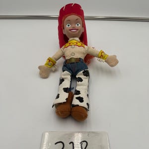 May include: A small Jessie doll from the Toy Story franchise. The doll has a red hat, a tan shirt with yellow accents, blue jeans, and cow-print chaps. The number "328" is written on a white label below the doll.