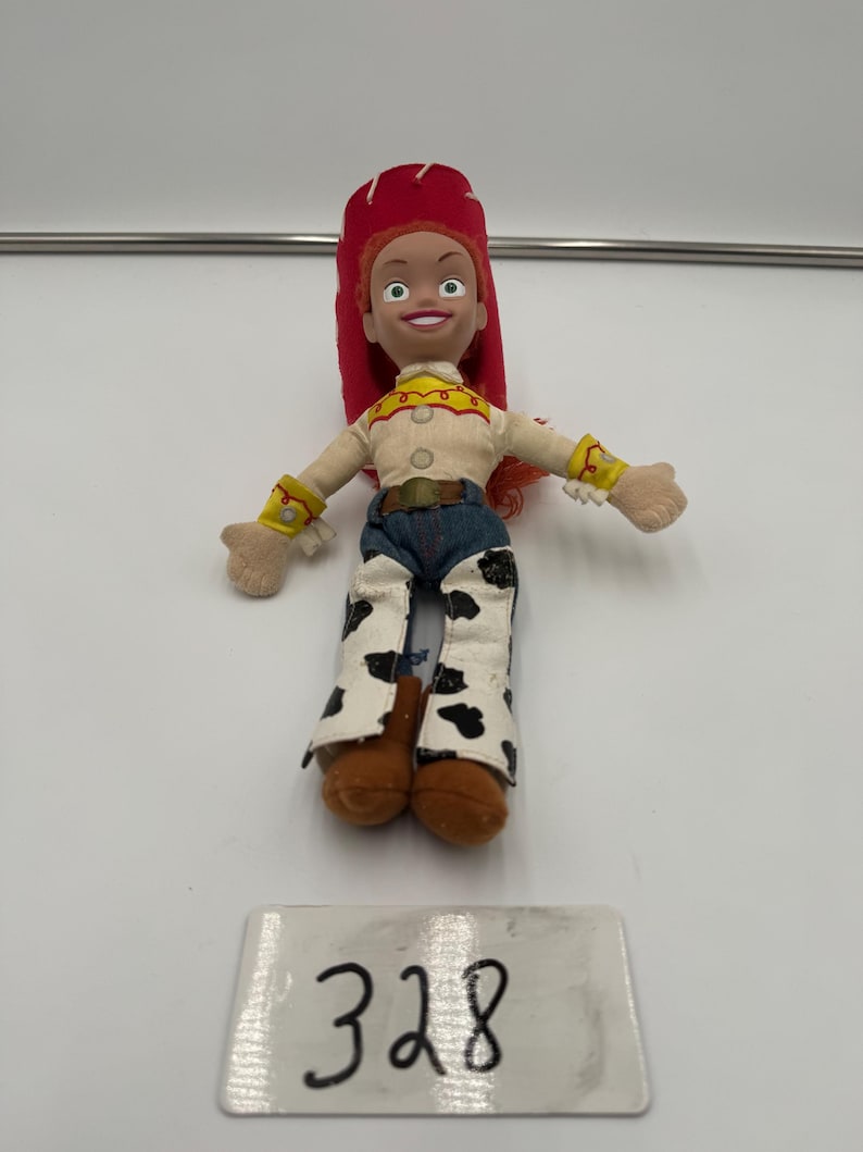 May include: A small Jessie doll from the Toy Story franchise. The doll has a red hat, a tan shirt with yellow accents, blue jeans, and cow-print chaps. The number "328" is written on a white label below the doll.