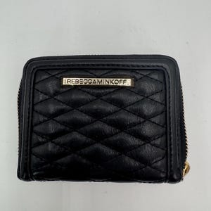 Puede incluir: Cartera de cuero negro acolchada con cierre de cremallera. La cartera presenta un patr&oacute;n de diamantes y una peque&ntilde;a placa met&aacute;lica rectangular con la marca "REBECCAMINKOFF". El tirador de la cremallera es dorado.