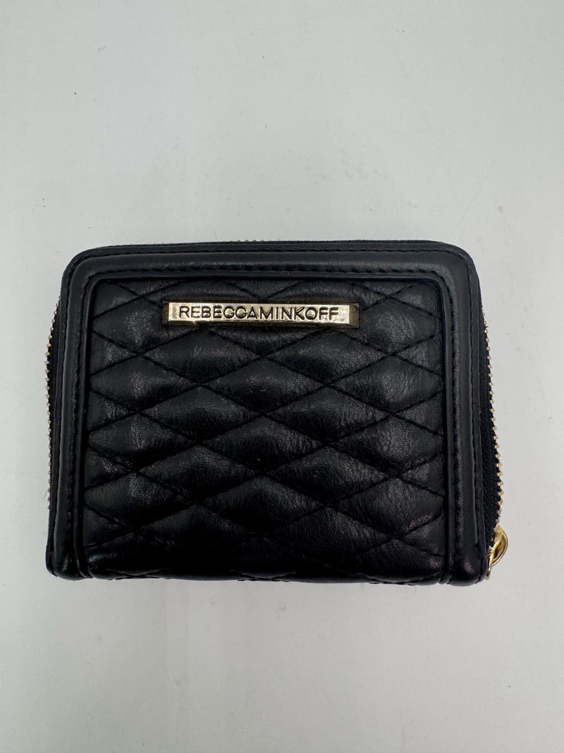 Puede incluir: Cartera de cuero negro acolchada con cierre de cremallera. La cartera presenta un patr&oacute;n de diamantes y una peque&ntilde;a placa met&aacute;lica rectangular con la marca "REBECCAMINKOFF". El tirador de la cremallera es dorado.