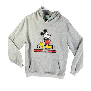Puede incluir: Sudadera con capucha gris con un gr&aacute;fico de Mickey Mouse y la palabra "GUCCI" en rojo y dorado. La sudadera tiene un bolsillo delantero y una capucha con cord&oacute;n. El logotipo de Disney es visible en la capucha.