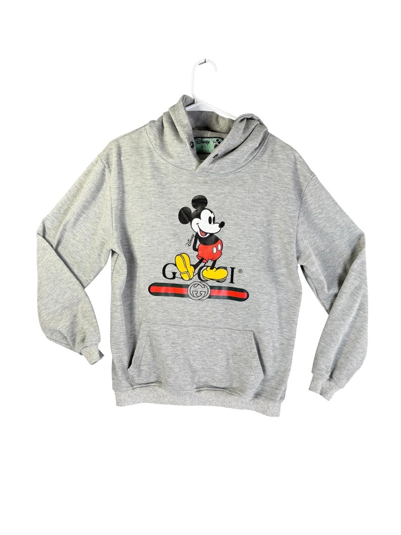 Puede incluir: Sudadera con capucha gris con un gr&aacute;fico de Mickey Mouse y la palabra "GUCCI" en rojo y dorado. La sudadera tiene un bolsillo delantero y una capucha con cord&oacute;n. El logotipo de Disney es visible en la capucha.