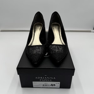 K&ouml;nnte beinhalten: Ein Paar schwarze Adriana Papell Pumps mit spitzer Zehenpartie und floralem Spitzendesign. Die Schuhe werden auf einer schwarzen Schuhschachtel pr&auml;sentiert. Auf der Schachtel sind der Markenname und die Schuhgr&ouml;&szlig;eninformationen aufgedruckt.