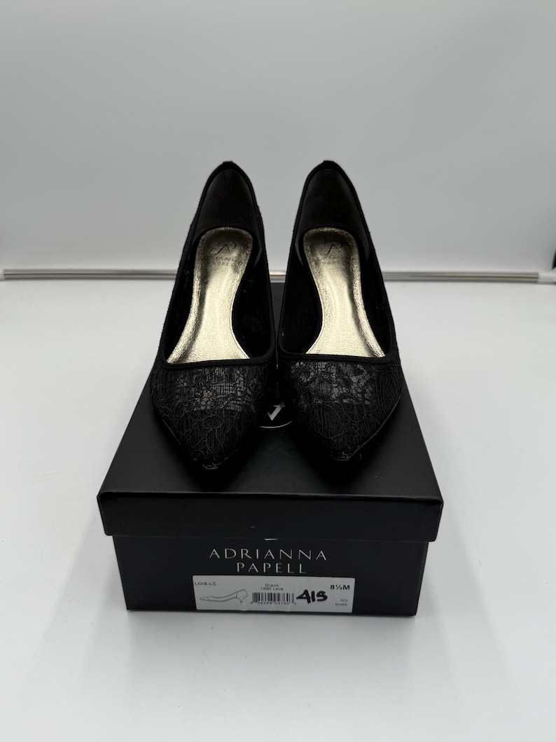 K&ouml;nnte beinhalten: Ein Paar schwarze Adriana Papell Pumps mit spitzer Zehenpartie und floralem Spitzendesign. Die Schuhe werden auf einer schwarzen Schuhschachtel pr&auml;sentiert. Auf der Schachtel sind der Markenname und die Schuhgr&ouml;&szlig;eninformationen aufgedruckt.