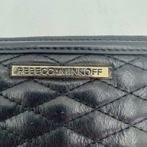 Puede incluir: Primer plano de una cartera de cuero negro acolchado. La cartera presenta un patr&oacute;n de diamantes y una placa rectangular dorada con el nombre de la marca "REBECCAMINKOFF". El cuero parece ligeramente texturizado.