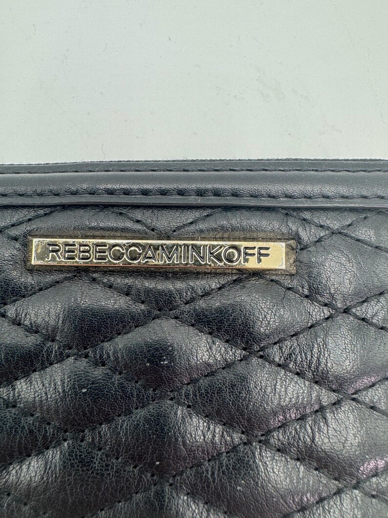 Puede incluir: Primer plano de una cartera de cuero negro acolchado. La cartera presenta un patr&oacute;n de diamantes y una placa rectangular dorada con el nombre de la marca "REBECCAMINKOFF". El cuero parece ligeramente texturizado.