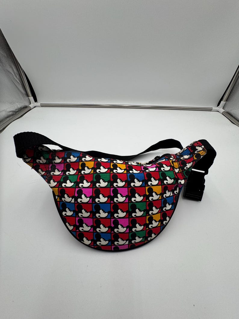 May include: A waist bag featuring a repeating grid pattern of Mickey Mouse silhouettes in black against colorful squares of red, yellow, green, blue, and pink. The bag includes a black adjustable strap with a black plastic side-release buckle.