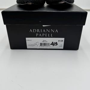 K&ouml;nnte beinhalten: Schwarze Spitzenschuhe von Adrianna Papell, Gr&ouml;&szlig;e 8 1/2 M, auf einer schwarzen Schachtel. Die Schachtel zeigt den Markennamen und Stilinformationen. Die Schuhe haben eine runde Spitze und einen niedrigen Absatz.