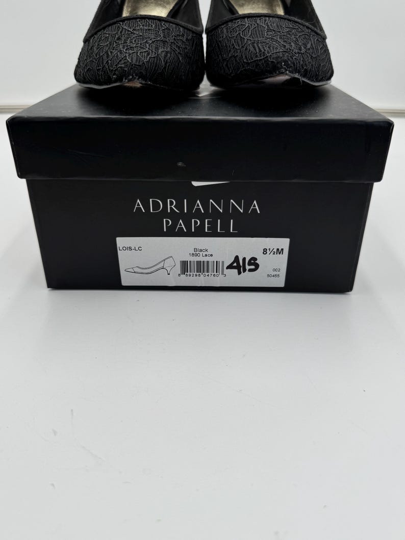 K&ouml;nnte beinhalten: Schwarze Spitzenschuhe von Adrianna Papell, Gr&ouml;&szlig;e 8 1/2 M, auf einer schwarzen Schachtel. Die Schachtel zeigt den Markennamen und Stilinformationen. Die Schuhe haben eine runde Spitze und einen niedrigen Absatz.