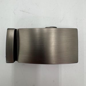 May include: A rectangular, brushed metal belt buckle in a gunmetal gray finish. The buckle has a simple, modern design with a smooth, horizontal texture. The buckle is designed to be used with a belt.