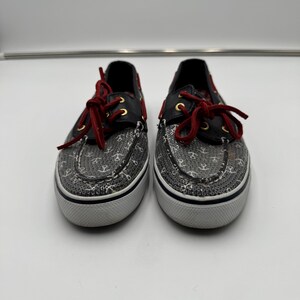 May include: A pair of boat shoes with a gray anchor pattern on the toe and sides. The shoes have black leather accents, red laces, and gold eyelets. The soles are white with a black stripe.