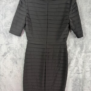 K&ouml;nnte beinhalten: Ein schwarzes, knielanges Kleid mit einem strukturierten, horizontal gefalteten Design. Das Kleid hat kurze &Auml;rmel und einen Rundhalsausschnitt. Es h&auml;ngt an einem wei&szlig;en Kleiderb&uuml;gel vor grauem Hintergrund.