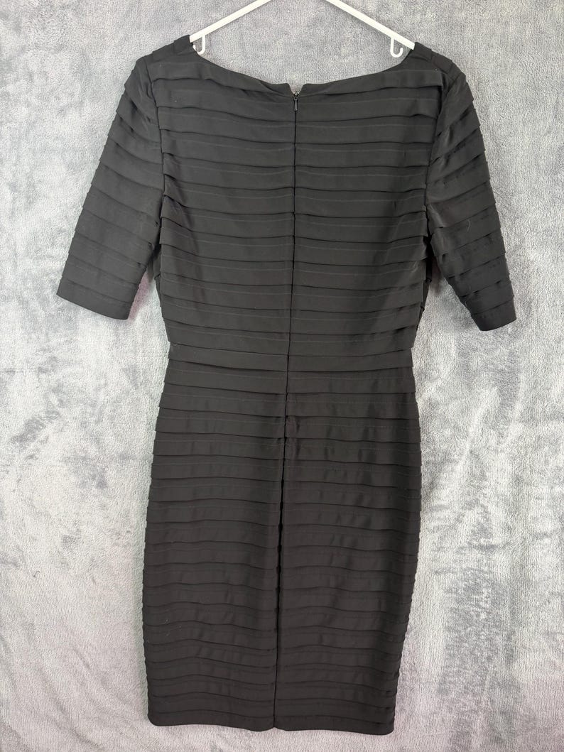 K&ouml;nnte beinhalten: Ein schwarzes, knielanges Kleid mit einem strukturierten, horizontal gefalteten Design. Das Kleid hat kurze &Auml;rmel und einen Rundhalsausschnitt. Es h&auml;ngt an einem wei&szlig;en Kleiderb&uuml;gel vor grauem Hintergrund.