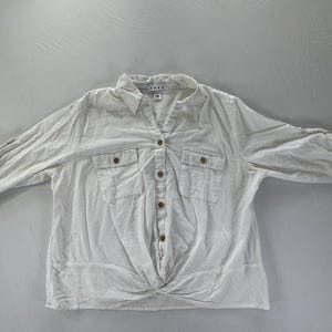 May include: A white button-down shirt with a V-neck collar, two chest pockets, and rolled-up sleeves. The shirt has a knotted detail at the waist and is made of a lightweight fabric.