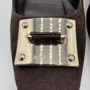 May include: Close-up of a brown suede shoe with a silver metal accent. The silver accent has a rectangular shape with rounded corners and a dark brown leather strap. The shoe has a pointed toe.