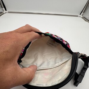 May include: A black waist bag with a colorful Mickey Mouse pattern on the exterior and a white interior lining. The bag features a black zipper closure, a black adjustable strap with a plastic buckle, and a small white tag inside with Disney branding.