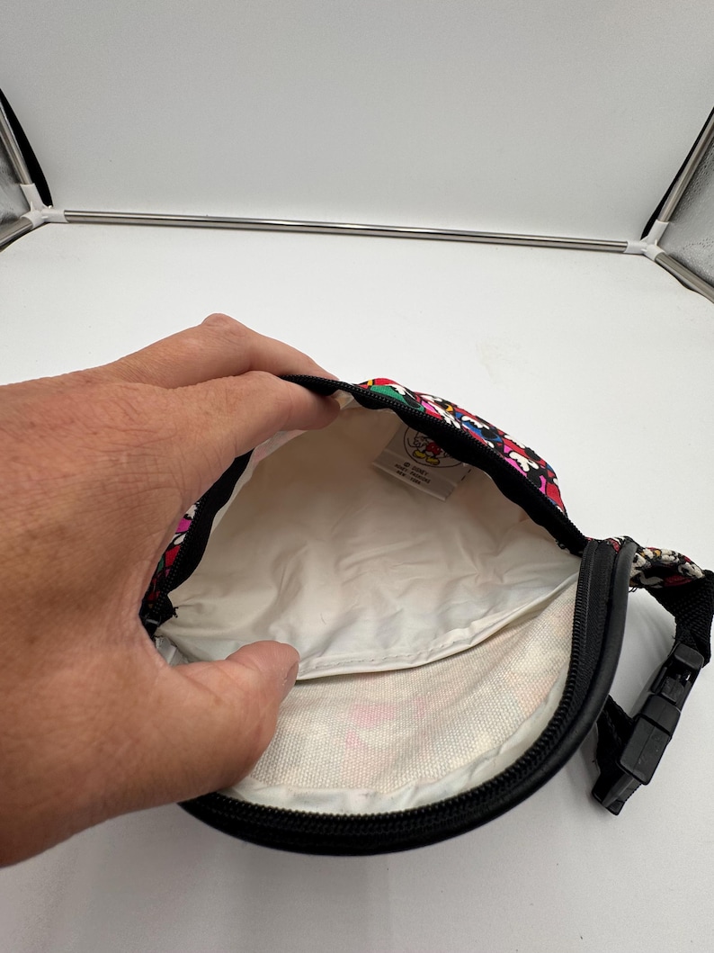 May include: A black waist bag with a colorful Mickey Mouse pattern on the exterior and a white interior lining. The bag features a black zipper closure, a black adjustable strap with a plastic buckle, and a small white tag inside with Disney branding.