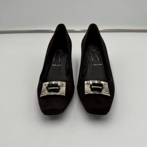 May include: A pair of dark brown low-heeled shoes. Each shoe has a square toe and a silver-colored buckle. The shoes appear to be made of a textured material. The background is plain white.