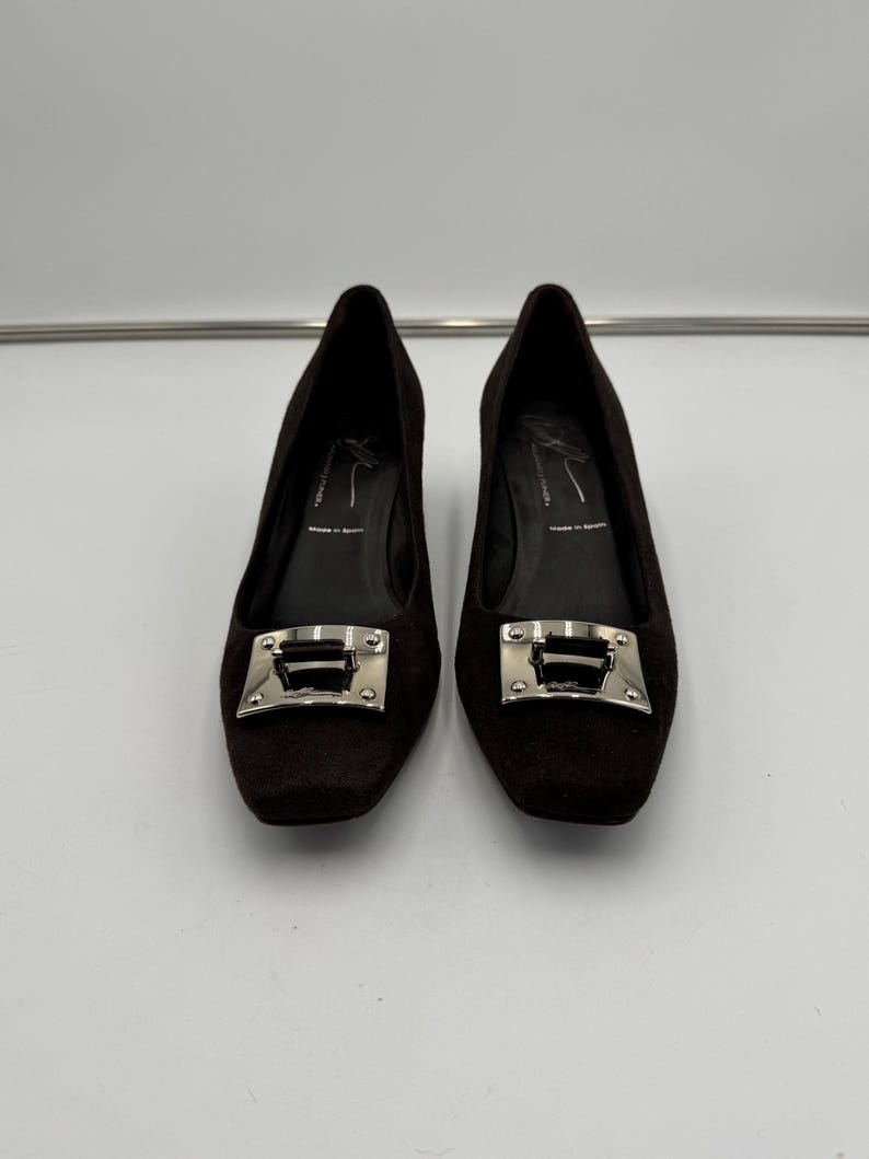 May include: A pair of dark brown low-heeled shoes. Each shoe has a square toe and a silver-colored buckle. The shoes appear to be made of a textured material. The background is plain white.