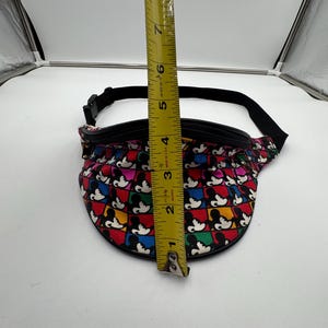 May include: A fanny pack featuring a repeating grid pattern of Mickey Mouse silhouettes in red, blue, yellow, and black squares. A yellow measuring tape is placed vertically over the center, showing the bag height is approximately 5.5 inches.