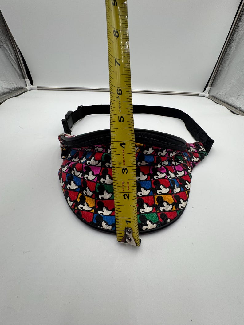 May include: A fanny pack featuring a repeating grid pattern of Mickey Mouse silhouettes in red, blue, yellow, and black squares. A yellow measuring tape is placed vertically over the center, showing the bag height is approximately 5.5 inches.