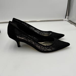 K&ouml;nnte beinhalten: Ein Paar schwarze Pumps aus Spitze mit spitzer Zehenpartie und niedrigen Abs&auml;tzen. Die Schuhe haben ein zartes florales Spitzenmuster und eine klassische Silhouette.