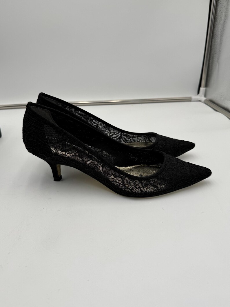 K&ouml;nnte beinhalten: Ein Paar schwarze Pumps aus Spitze mit spitzer Zehenpartie und niedrigen Abs&auml;tzen. Die Schuhe haben ein zartes florales Spitzenmuster und eine klassische Silhouette.