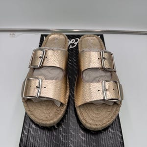 May include: Gold-colored sandals with two adjustable straps and silver buckles. The sandals have a woven, espadrille-style sole and are displayed on a black and white patterned box. The sandals are new.