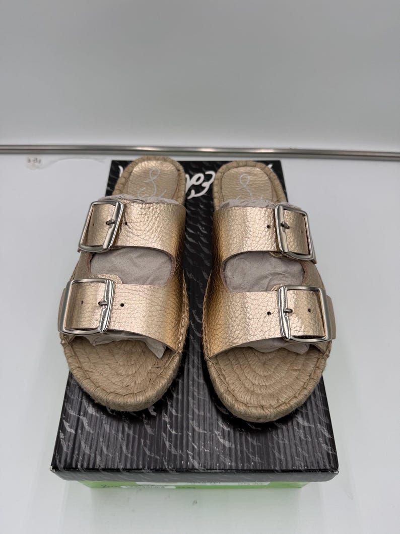 May include: Gold-colored sandals with two adjustable straps and silver buckles. The sandals have a woven, espadrille-style sole and are displayed on a black and white patterned box. The sandals are new.