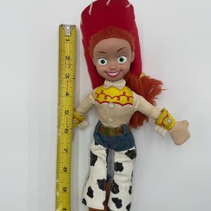 May include: A Jessie doll from the Toy Story franchise. The doll has red hair, a red hat, and a yellow shirt with a white top. She wears blue jeans with black and white cow print chaps. The doll is approximately 10 inches tall.