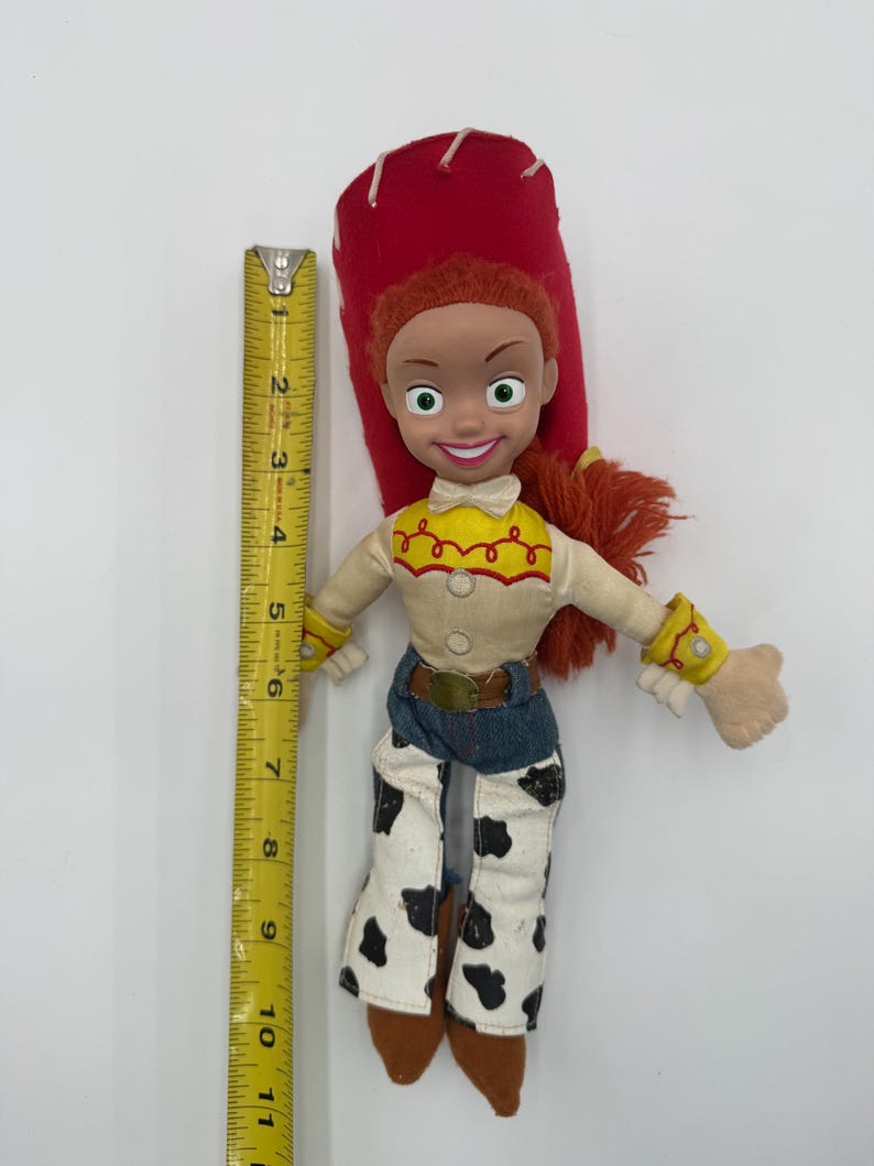 May include: A Jessie doll from the Toy Story franchise. The doll has red hair, a red hat, and a yellow shirt with a white top. She wears blue jeans with black and white cow print chaps. The doll is approximately 10 inches tall.