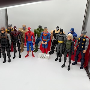 May include: A collection of superhero action figures, including Spider-Man, Superman, Thor, and Batman. The figures are brightly colored and detailed, with various costumes and accessories. A small white tag with the number 337 is visible.