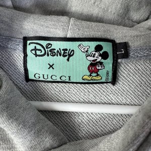 Puede incluir: Primer plano de una sudadera con capucha gris con una etiqueta de Disney x Gucci. La etiqueta es rectangular con un fondo verde claro y presenta el logotipo de Disney, el logotipo de Gucci y un gr&aacute;fico de Mickey Mouse.