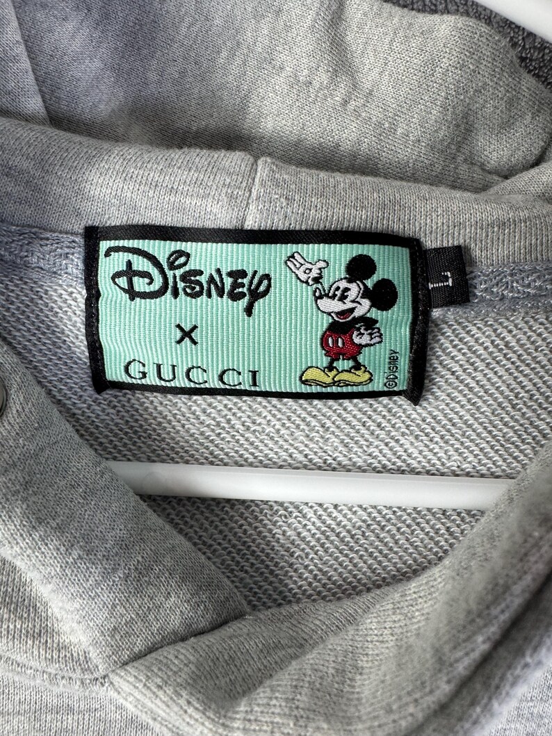 Puede incluir: Primer plano de una sudadera con capucha gris con una etiqueta de Disney x Gucci. La etiqueta es rectangular con un fondo verde claro y presenta el logotipo de Disney, el logotipo de Gucci y un gr&aacute;fico de Mickey Mouse.