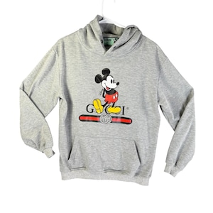 Puede incluir: Sudadera con capucha gris con un gr&aacute;fico de Mickey Mouse y la palabra "GUCCI" en rojo y dorado. La sudadera tiene un bolsillo delantero y una capucha con cord&oacute;n. El logotipo de Disney es visible en la capucha.
