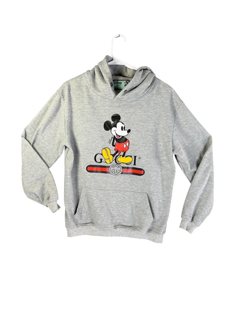 Puede incluir: Sudadera con capucha gris con un gr&aacute;fico de Mickey Mouse y la palabra "GUCCI" en rojo y dorado. La sudadera tiene un bolsillo delantero y una capucha con cord&oacute;n. El logotipo de Disney es visible en la capucha.